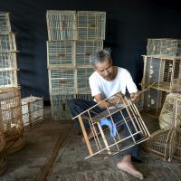 Senior focused craftsman with barefoot adding bamboo sticks in wooden carcass of bird cage while working in workshop in daytime